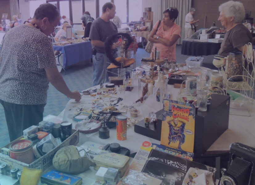 Visitors browsing antiques and collectibles at vendor tables during a busy indoor antiques show and expo.
