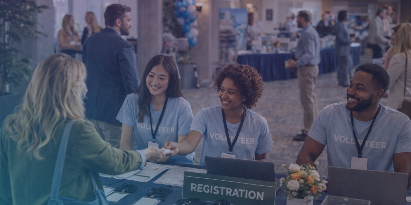 Volunteers assisting an attendee at a nonprofit event registration desk while guests network in the background at a community conference.