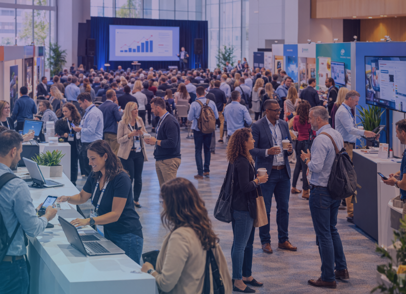 Busy corporate user conference in a large convention hall with attendees networking, visiting exhibitor booths, and listening to a keynote presentation while organizers manage registration at a check-in desk.