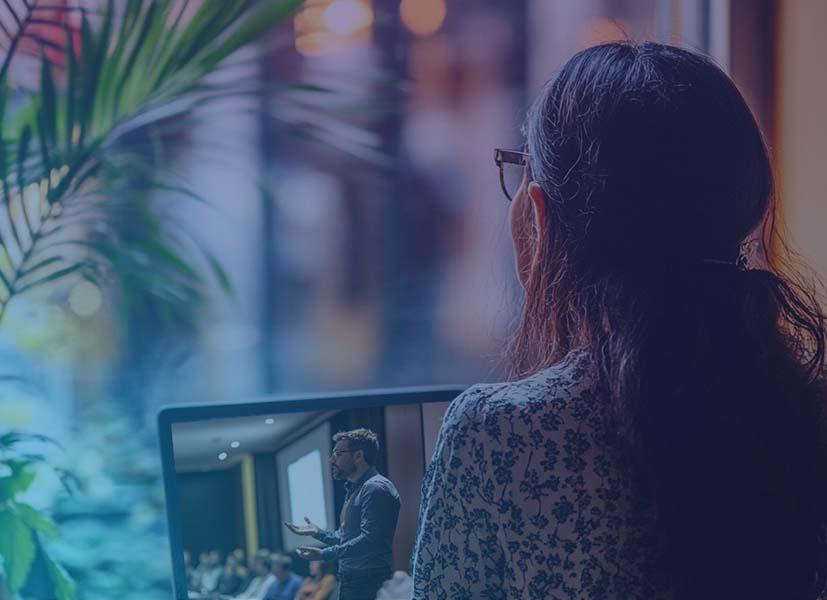 Image of a female hybrid conference participant sitting at a desk watching a conference speaker presenting on a laptop.