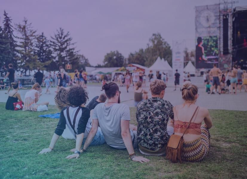 Attendees sitting on grass at an outdoor community festival with vendor tents, live stage, and crowds in the background.