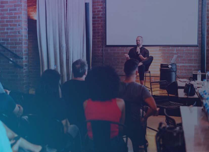 Image of a speaker on stage presenting a topic during a live seminar event in front of a seated audience.